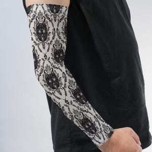 May include: An arm sleeve featuring a cream-colored background with a repeating black damask pattern that incorporates stylized cat faces. The sleeve extends from the upper arm to the wrist, displaying a detailed, symmetrical graphic design.