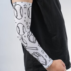 May include: A white compression arm sleeve featuring a repeating black line art pattern of baseballs, baseball gloves, bats, and small stars. The sleeve is worn on the forearm and extends toward the bicep, covering the skin with a sports-themed design.