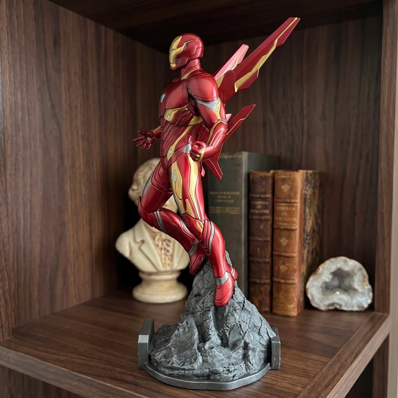 May include: A red and gold Iron Man action figure in a dynamic flying pose, mounted on a textured gray rocky base. The figure features mechanical armor plating, a sleek helmet, and prominent wing-like structures extending from the back.
