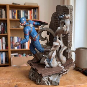 May include: A Captain America action figure in a blue suit with a brown belt and gloves, running forward. The figure is mounted on a base featuring a brick wall, wooden planks, and swirling dust effects, with a circular shield attached to the back.