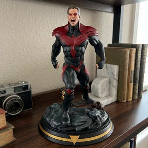 Op de afbeelding: Een gedetailleerd verzamelfiguur van een gespierd mannelijk personage, gekleed in een zwart en rood pak met een opvallend rood adelaarembleem op de borst. Het figuur heeft een open mond, rode ogen en staat op een rotsachtige basis.
