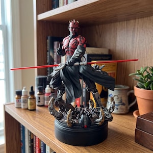 Op de afbeelding: Een gedetailleerd beeldje van een rood en zwart gehoornd personage met gezichtstatoeages, donkere gewaden en metalen beenharnas. Het figuur houdt een lang, dubbelzijdig rood lichtzwaard horizontaal vast op een donkere, getextureerde ronde voet.