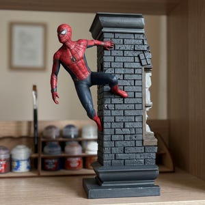 May include: A detailed figurine of the superhero Spider-Man in a red and black suit, depicted in a dynamic climbing pose against a vertical, dark grey textured brick wall structure with a partial window frame detail on the side.