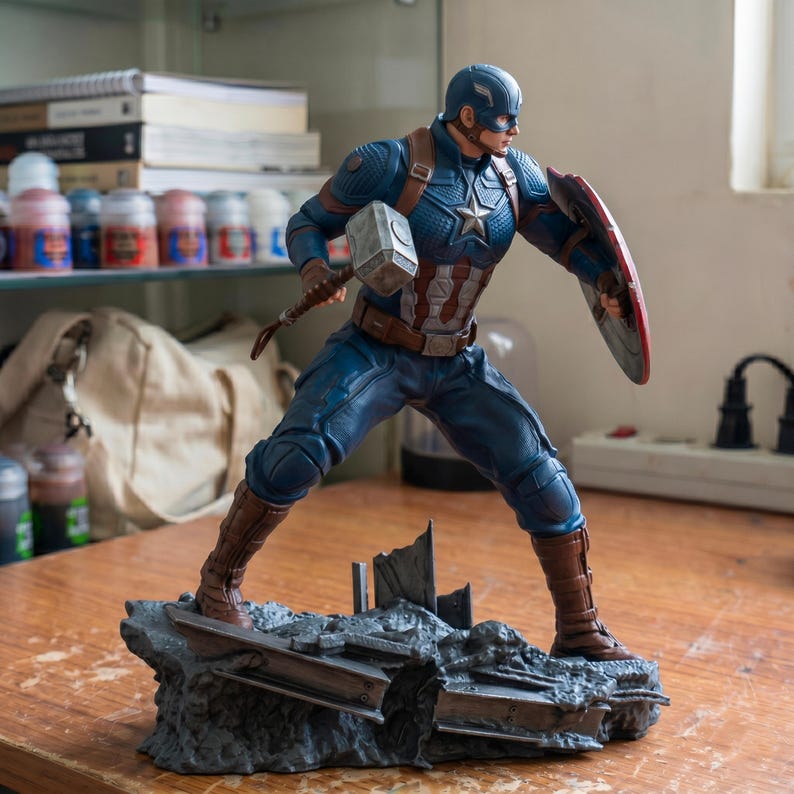 K&ouml;nnte beinhalten: Eine Captain America-Figur in dynamischer Pose, die einen Hammer und einen Schild h&auml;lt. Die Figur tr&auml;gt ein blaues, wei&szlig;es und rotes Kost&uuml;m mit einem Sternemblem auf der Brust. Der Sockel ist ein detailliertes, graues, felsiges Gel&auml;nde.