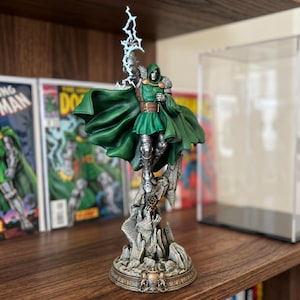 May include: A detailed figurine of Doctor Doom, a comic book character, stands on a rocky base. The figure is clad in a green tunic and cape, with silver armor and a metal mask. He holds a blue lightning bolt. Comic books are in the background.