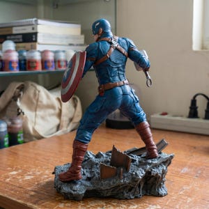 K&ouml;nnte beinhalten: Eine Captain America-Figur in dynamischer Pose, die ihren Schild und einen Hammer h&auml;lt. Die Figur tr&auml;gt ein blaues und braunes Kost&uuml;m und steht auf einem felsigen Sockel. Der Schild ist rot, wei&szlig; und blau.