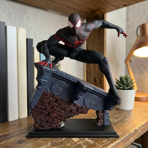Pode incluir: Uma figura de ação do Spider-Man Miles Morales em pose agachada, com um traje preto com padrões de teia vermelhos e um emblema de aranha vermelho no peito. A figura está montada numa base que imita uma cornija de tijolo desgastada.