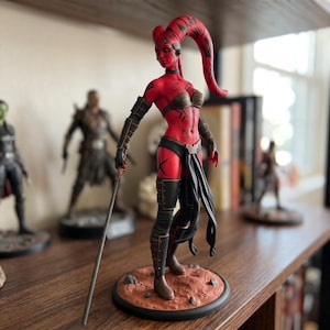 May include: A red-skinned humanoid character figurine with long head-tails, wearing a dark metallic bikini top, black arm guards, and a black loincloth. The figure stands on a textured reddish-brown base and holds a long, thin silver-coloured staff.