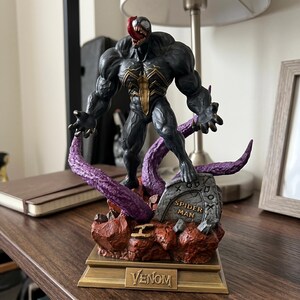 May include: A detailed Venom figurine featuring a muscular black creature with a gold spider emblem, a long red tongue, and sharp teeth. It stands on a rocky base with purple tentacles and a tombstone marked SPIDER MAN, set on a gold base labelled VENOM.