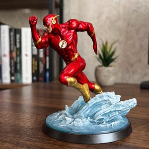 Könnte beinhalten: Eine dynamische Figur von The Flash in Laufpose, mit einem metallisch roten Anzug mit goldenen Akzenten und einem kreisförmigen Blitz-Emblem auf der Brust. Die Figur ist über einem durchscheinenden blauen Sockel positioniert, der wie Wasser geformt ist.