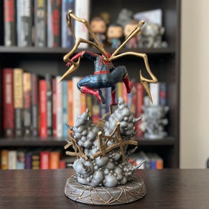 Op de afbeelding: Een gedetailleerd actiefiguur van een superheld in een rood en blauw pak, met vier gouden mechanische spinnenpoten op de rug. Het figuur bevindt zich in een zwevende pose boven een basis van grijze rookwolken en gouden structurele brokstukken.