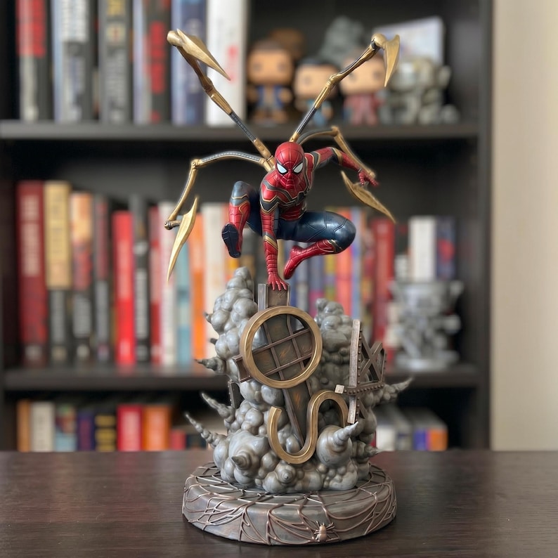 Op de afbeelding: Een Spider-Man actiefiguur in een rood en blauw pak met vier gouden mechanische spinnenpoten op de rug. Het figuur staat in een gehurkte houding op een grijze, wolkachtige basis met een cirkelvormige structuur en een platform met webpatroon.