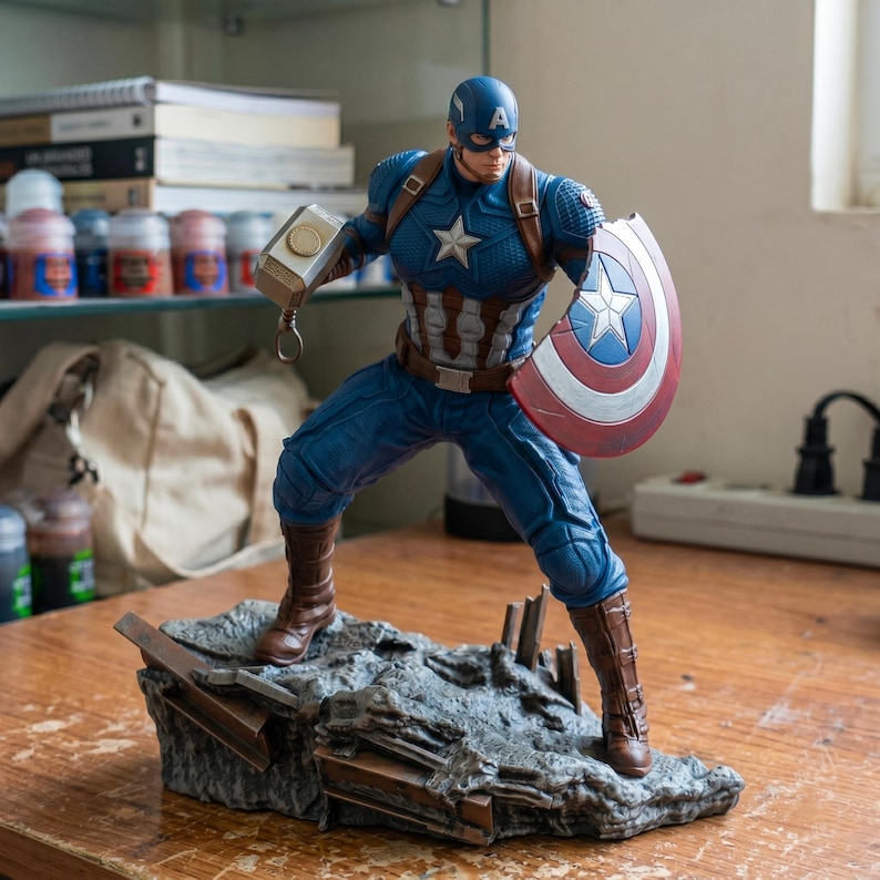 K&ouml;nnte beinhalten: Eine Captain America-Figur in dynamischer Pose, die einen Hammer und einen Schild h&auml;lt. Die Figur tr&auml;gt ein blau-wei&szlig;es Kost&uuml;m mit einem Sternemblem. Der Sockel ist ein graues, felsiges Gel&auml;nde, und der Schild hat ein rot-wei&szlig;-blaues Design.