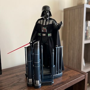 Puede incluir: Una figura de acción de Darth Vader de pie sobre una base de plataforma detallada. La figura lleva un casco, capa y traje negros con un panel de control en el pecho con botones iluminados. Una fina hoja de sable de luz roja sobresale.