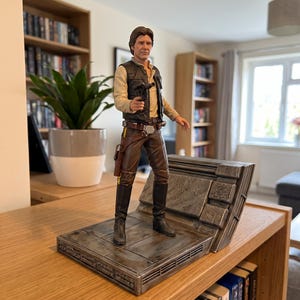 May include: A detailed Han Solo action figure standing on a textured, metallic-looking base. The figure wears a cream-coloured long-sleeve shirt, a black vest, brown trousers, and black boots, holding a small black blaster pistol in the right hand.