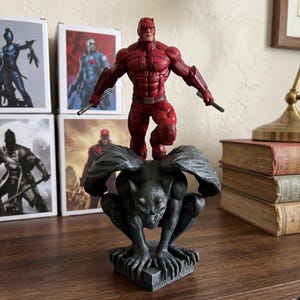 Puede incluir: Una figura de acción roja de Daredevil de pie sobre una base de gárgola de color gris oscuro con textura de piedra. La figura viste un traje rojo texturizado con máscara, cinturón y brazaletes, sosteniendo dos bastones plateados.