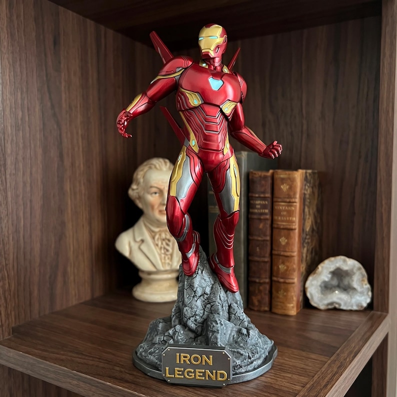 May include: A detailed Iron Man action figure standing on a rocky base with a plaque reading IRON LEGEND. The figure features a metallic red and gold suit with a light blue arc reactor on the chest, posed in a dynamic stance with wings extended.
