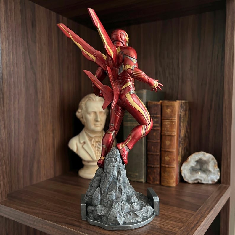 May include: A red and gold Iron Man action figure in a dynamic pose, featuring mechanical wings extending from the back. The figure is mounted on a textured gray rock-like base, showcasing intricate armor plating details and a sleek, metallic finish.