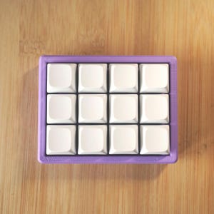 May include: A small, square keyboard in purple, featuring twelve white keycaps. The keyboard sits on a wooden surface. The keycaps are arranged in a 3x4 grid. The wooden surface has a light brown colour.