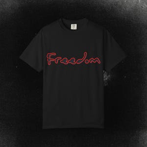 Puede incluir: Camiseta negra con la palabra "Freedom" en rojo, escrita en cursiva en el pecho. La camiseta tiene mangas cortas y cuello redondo. El fondo es una superficie oscura y texturizada.