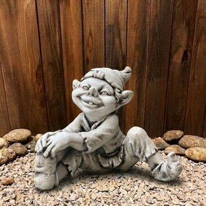 Puede incluir: Estatua de gnomo de jardín de hormigón gris con una cara sonriente, orejas puntiagudas y un sombrero. El gnomo está sentado con las piernas estiradas, las manos apoyadas en las piernas. La estatua está sobre un lecho de pequeñas rocas, con una valla de madera al fondo.