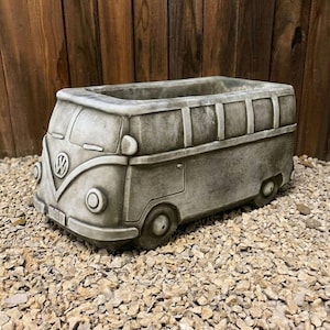 Puede incluir: Una jardinera de hormigón gris con forma de furgoneta Volkswagen vintage. La jardinera tiene una abertura rectangular para plantas, con detalles como el logotipo de VW, los faros y las ruedas. Se encuentra sobre un lecho de pequeñas piedras, con un fondo de madera.