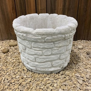 May include: A light gray, stone-like planter with a textured, brick-like design. The planter has a wide, open top and is sitting on a bed of small, tan pebbles. The background is a wooden fence.