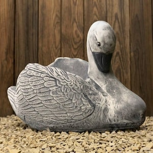 May include: A grey swan planter with detailed feather textures. The swan has a black beak and is resting on small, light-coloured stones. The background is a wooden wall.