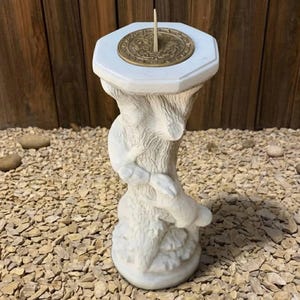 May include: A white sundial with a bronze-colored face and gnomon. The sundial is supported by a sculpted base with a tree-like design. The sundial is a decorative garden accent.