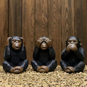 May include: Three monkey figurines in a row, each in a different pose: covering ears, eyes, and mouth. The monkeys are black with brown accents, sitting on a bed of small rocks, against a wooden background.