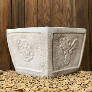 May include: A square, off-white planter with embossed floral designs. The planter has a textured surface and is set on a bed of small, tan stones. The background is a wooden wall.