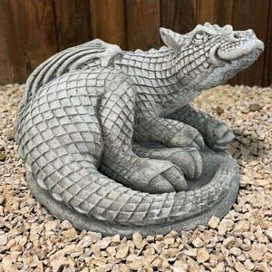 Puede incluir: Estatua de dragón gris con escamas texturizadas, alas y cola enroscada. El dragón descansa sobre una base circular, rodeado de pequeños guijarros. La escultura tiene una cara y garras detalladas.
