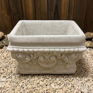 Può includere: Una fioriera quadrata in cemento grigio chiaro con dettagli floreali ornati. La fioriera ha un bordo largo e poggia su un letto di piccole pietre chiare. Lo sfondo presenta assi di legno e alcune rocce più grandi.