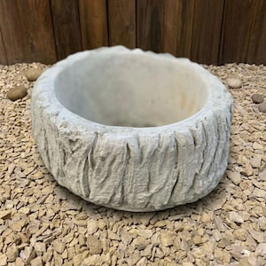 May include: A round, light gray stone planter with a textured, bark-like exterior. The planter sits on a bed of small, light-colored gravel. The interior is smooth and open, ready for planting.