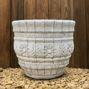 Può includere: Un vaso per piante in cemento grigio chiaro di forma cilindrica, caratterizzato da una superficie strutturata con pannelli verticali e un motivo floreale in rilievo ripetuto al centro. Il bordo ha un design smerlato con finitura ruvida.