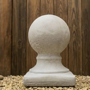 Könnte beinhalten: Ein hellgraues Gartenkugel-Zierstück aus Beton mit strukturierter Oberfläche. Das Zierstück hat eine runde Oberseite, einen dekorativen Sockel und einen quadratischen Boden. Der Hintergrund ist eine Holzwand.