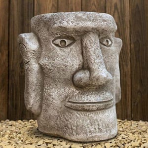 May include: A weathered, grey stone head sculpture with a large nose, closed eyes, and a smiling mouth. The sculpture has a textured surface, likely a decorative garden ornament or planter.