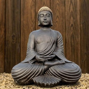 Può includere: Una statua di Buddha grigio scuro in posizione di meditazione seduta. La statua ha un'espressione serena, occhi chiusi e un copricapo dettagliato. La figura è drappeggiata in vesti fluenti, con le mani appoggiate in grembo. La statua è posta su un letto di piccole pietre.