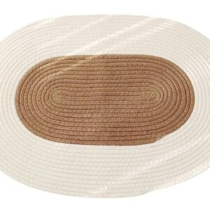 Può includere: Un tappeto intrecciato di forma ovale con una sezione centrale in fibre color beige naturale circondata da un ampio bordo in materiale intrecciato bianco sporco. La struttura è composta da anelli concentrici di corda spessa e testurizzata.