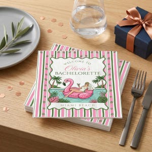Puede incluir: Una pila de servilletas decorativas con un borde a rayas rosas y verdes. Las servilletas presentan un diseño de flotador de flamenco con el texto "Welcome to Olivia's Bachelorette" y "Miami Beach". También se ven un tenedor, un cuchillo y una caja de regalo.