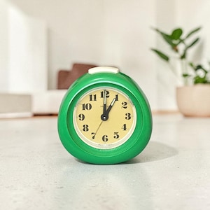 Peut inclure: Un réveil vert vintage avec un cadran crème et des chiffres noirs. La montre a un design arrondi et un bouton blanc sur le dessus. Les aiguilles indiquent environ 12h05. Une petite plante est visible en arrière-plan.