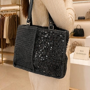 May include: Black tote bag with a woven texture and sequin embellishments. The bag has a rectangular shape with a black leather handle. The bag is part of the Yeni Koleksiyon.