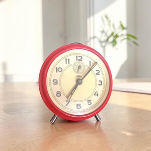 Può includere: Una sveglia analogica rossa con quadrante color crema e lancette nere. L'orologio ha una forma rotonda e poggia su due piedini argentati. I numeri sono neri e il quadrante ha un piccolo quadrante nella parte superiore.