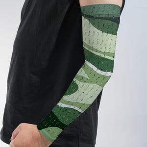May include: An arm sleeve featuring a repeating pattern of small, dark green figures in various athletic poses against a background of wavy, layered bands in shades of light sage, olive, and dark forest green, worn with a plain black t-shirt.