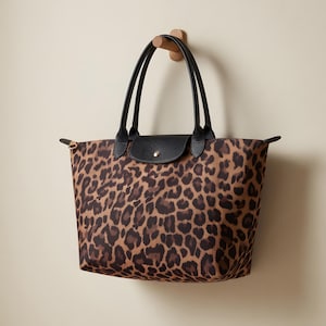 May include: A tote bag with a leopard print pattern. The bag has black leather handles and a black flap with a gold-colored button closure. The bag is hanging on a wooden hook.