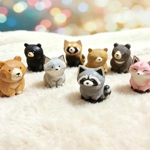 Può includere: Una collezione di nove figurine di animali in legno intagliato, tra cui orsi, una volpe, un procione e un panda rosso. Le figurine sono in vari colori, come marrone, nero, grigio e rosa. Gli animali sono esposti su una superficie bianca e soffice.