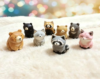 Könnte beinhalten: Eine Sammlung von neun geschnitzten Holzfiguren von Tieren, darunter Bären, ein Fuchs, ein Waschbär und ein Roter Panda. Die Figuren sind in verschiedenen Farben wie Braun, Schwarz, Grau und Rosa gehalten. Die Tiere sind auf einer weißen, flauschigen Oberfläche ausgestellt.