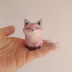 May include: A small, pink wooden fox figurine with black ears and eyes. The fox is carved with a smiling expression and has a white chest and belly. The figurine is approximately 7.6 cm tall.