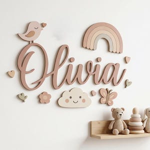 Może przedstawiać: Drewniany napis na ścianę z imieniem Olivia wykonany kursywą, ozdobiony tęczą, ptaszkiem, chmurką, motylem, kwiatami i sercami w stonowanych odcieniach różu, beżu i szałwiowej zieleni, umieszczony nad drewnianą półką z zabawkami.