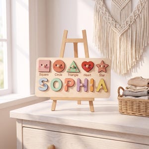 May include: A rectangular wooden puzzle board featuring the name SOPHIA in large, colorful letters and a row of five shapes above it: a square, circle, triangle, heart, and star. Each shape and letter includes a small wooden knob for grasping.